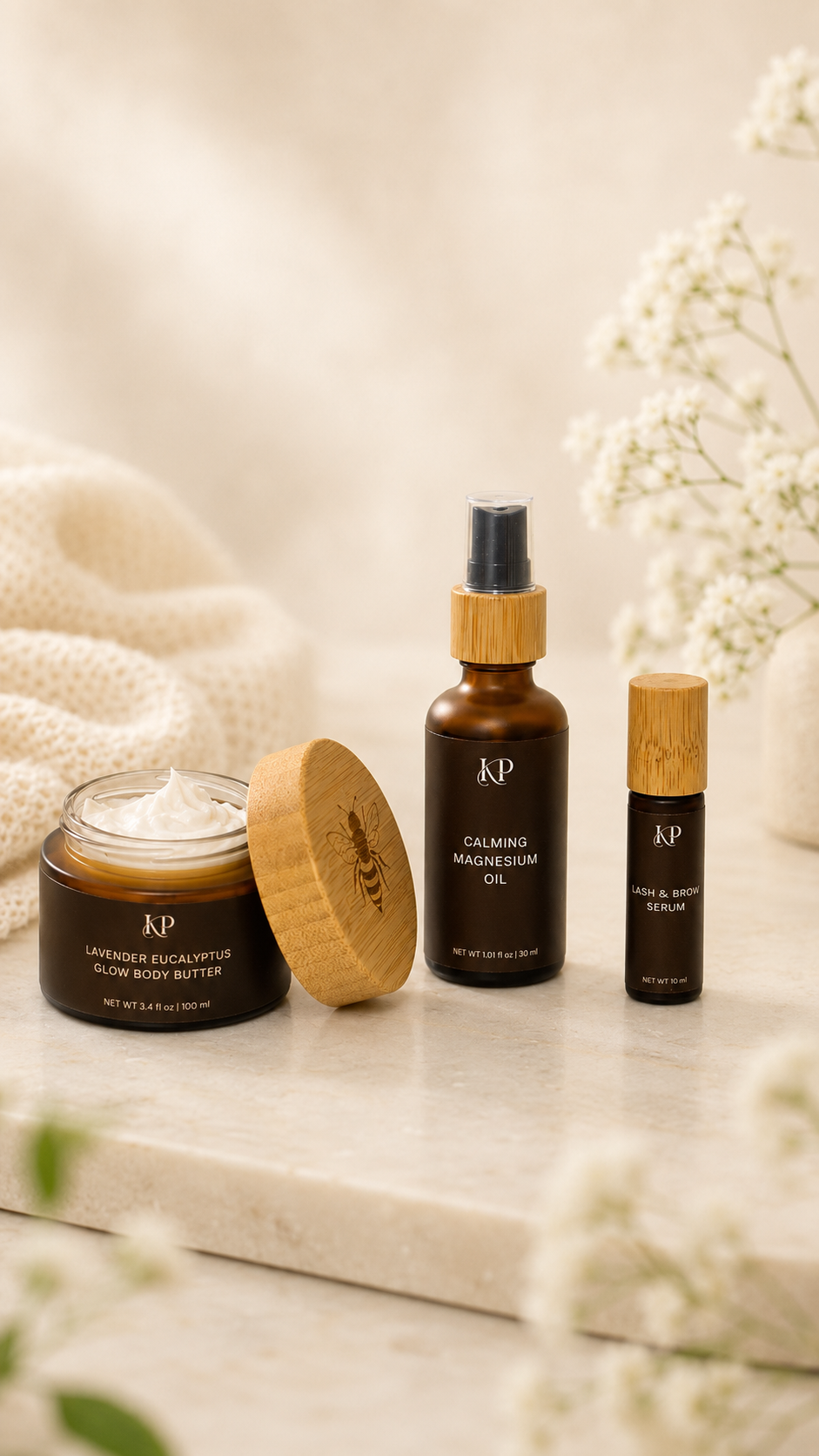 Three KP skincare products arranged on a light stone surface: a jar of lavender  glow body butter with its wooden lid beside it, a bottle of calming magnesium oil, and a small roll-on serum bottle, styled with soft cream fabric and white flowers.